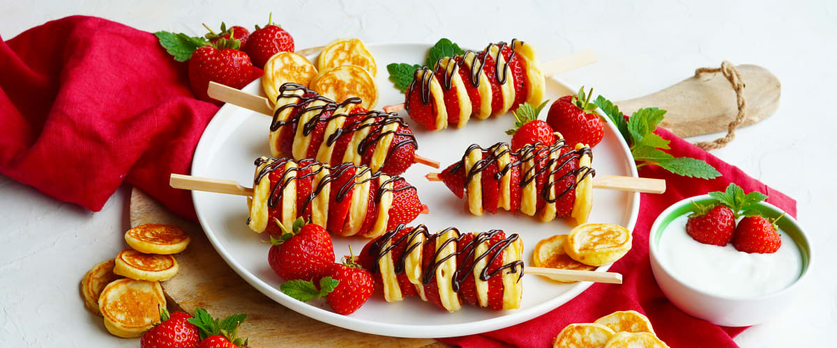 Palačinkové špízy s jahodami a čokoládou na talíři, vedle malých palačinek, jahod a misky s jogurtem