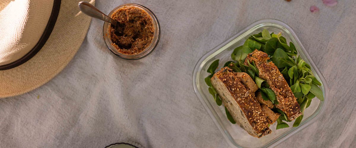 Sendvič s listy salátu, sklenice s pomazánkou, ořechy, klobouk a sklenice nápoje na piknikovém ubrusu