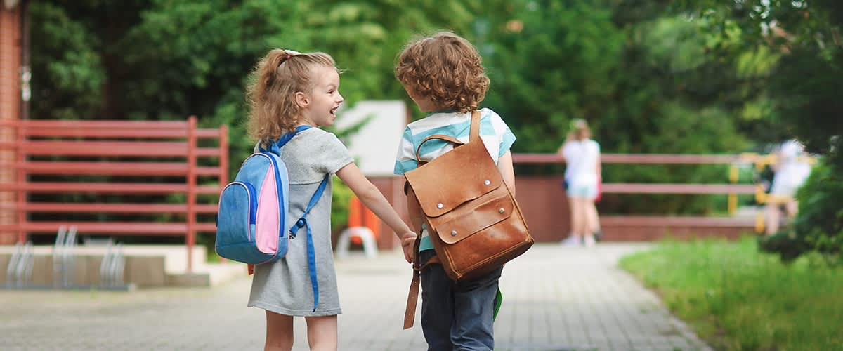Dvě děti s batohy držící se za ruce na chodníku venku