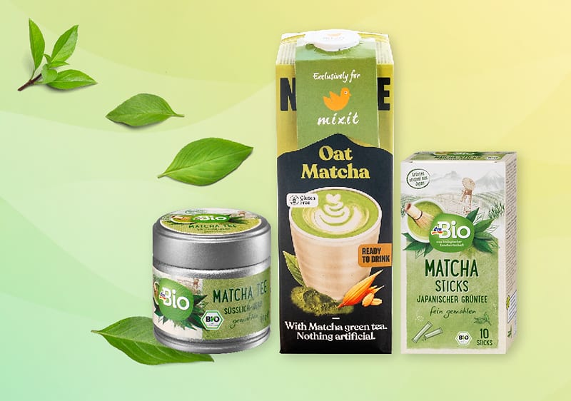 Produkty s matcha čajem: plechovka, krabice s nápojem, balení matcha tyčinek, zelené listy na pozadí