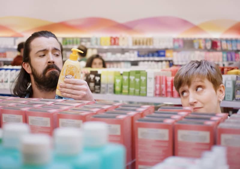 Lidé nakupují v obchodě s kosmetikou, drží lahvičku, regály plné produktů v pozadí