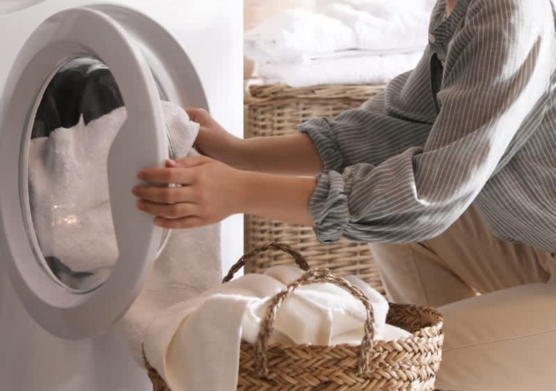 Žena vkládá bílé prádlo do pračky, vedle ní jsou proutěné koše na prádlo