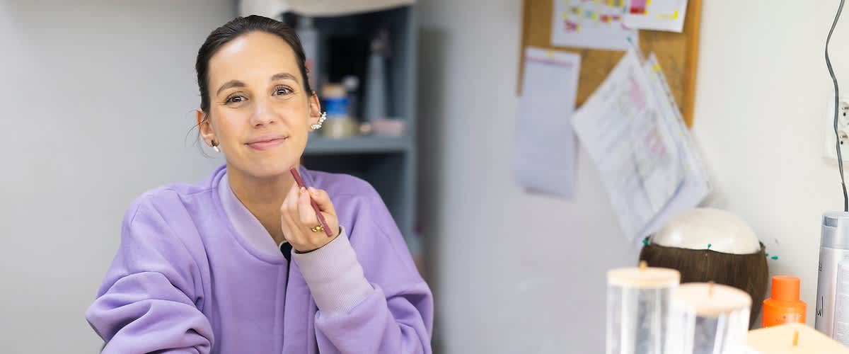 Žena v fialové mikině sedí u stolu s kosmetikou, rozmazaný obličej, zrcadlo a police v pozadí