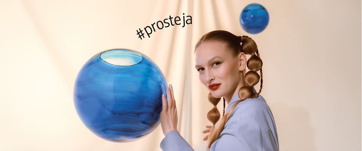 Žena s copánky drží modrou skleněnou kouli, v pozadí další koule a text #prosteja