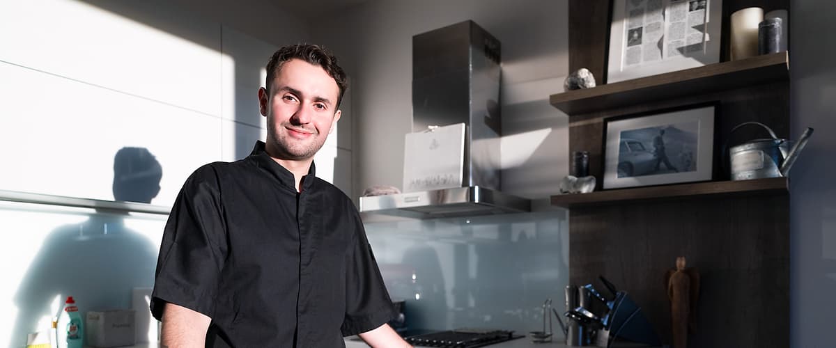 Osoba stojí v moderní kuchyni, na pultu jsou dezerty a pečivo.