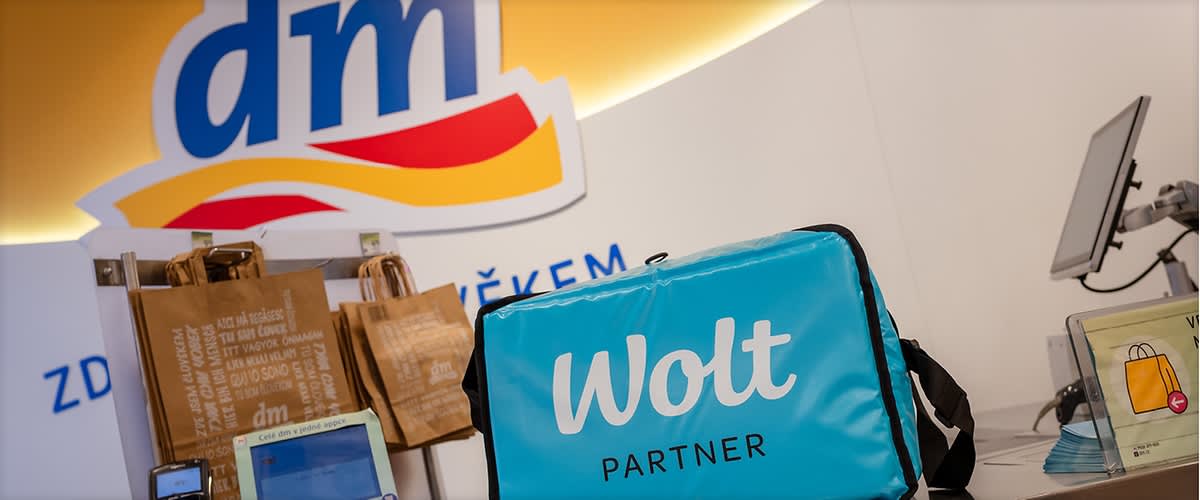 Tmavě modré logo dm nad pultem s nákupy a tyrkysovou Wolt doručovací taškou
