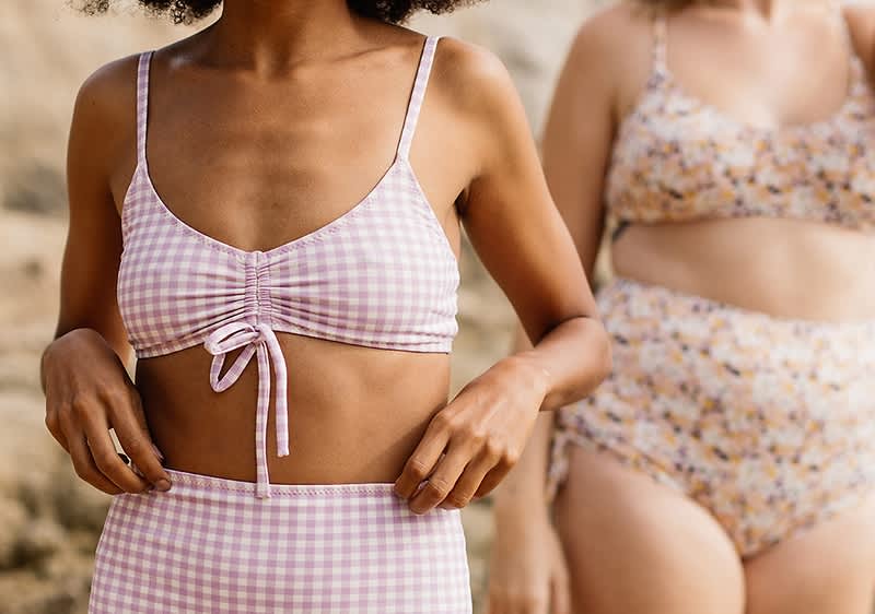Dvě ženy ve stylových plavkách stojící na pláži, jedna v kostkovaném a druhá v květovaném bikinovém setu