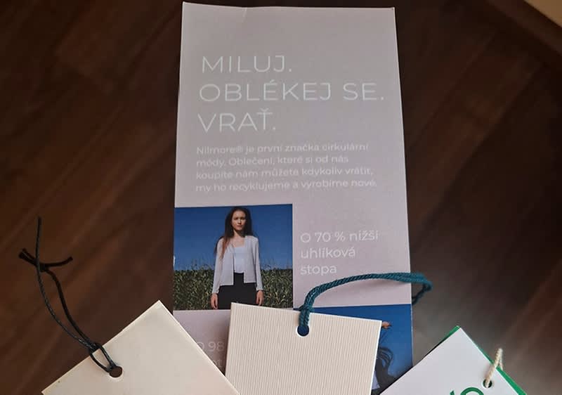 Svislý leták s nápisem „MILUJ. OBLEKEJ SE. VRAŤ.“ nad krátkým textem o návratnosti oblečení, uprostřed menší fotografie ženy v bílém saka na poli a vedle údaj „o 70 % nižší uhlíková stopa“, dole visí tři vzorníky se zavazovacími visačkami