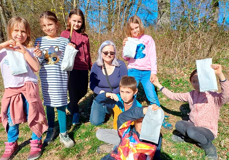 Skupina sedmi dětí a dospělé osoby venku na trávě u lesa, většina drží bílé papírové sáčky s nasbíranými předměty, všichni se usmívají a stojí nebo sedí na slunném dni
