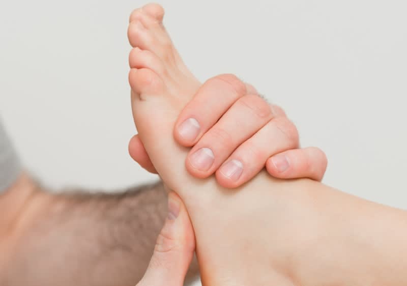 Dvě ruce drží a masírují nárt a chodidlo druhé osoby, vidět prsty a nehty, neutrální světlá pokožka, světlé pozadí