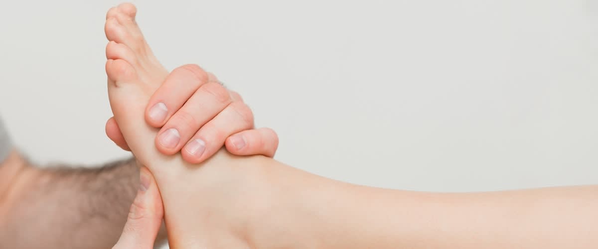 Dvě ruce drží a masírují nárt a chodidlo druhé osoby, vidět prsty a nehty, neutrální světlá pokožka, světlé pozadí