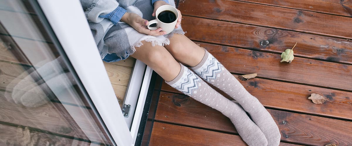 Žena sedí na parapetu otevřeného okna, přikrývená šedým plédem, drží bílý hrnek s černou kávou a má natáhnuté světle šedé až podkolenkové ponožky se vzorem, nohy vystrčené na dřevěné terase s několika opadanými listy
