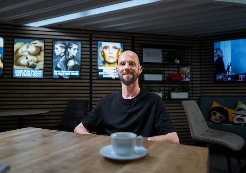 Muž sedí u dřevěného stolu v kavárně, usmívá se, má černé tričko a před ním stojí bílý šálek kávy na podšálku, v pozadí reklamní obrazovky a sedací nábytek