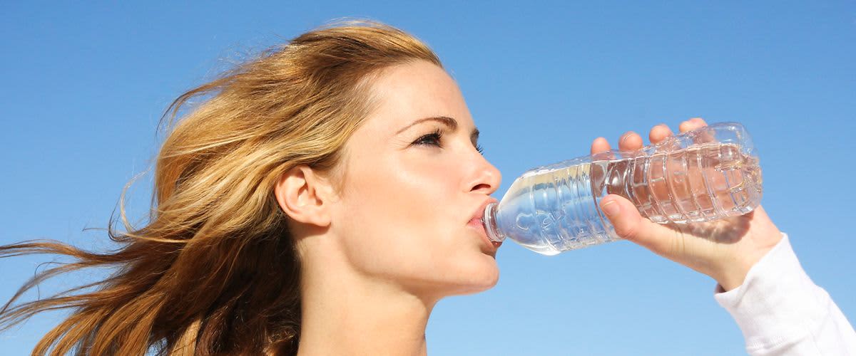 Žena pije vodu z plastové lahve na slunečném dni s modrou oblohou v pozadí