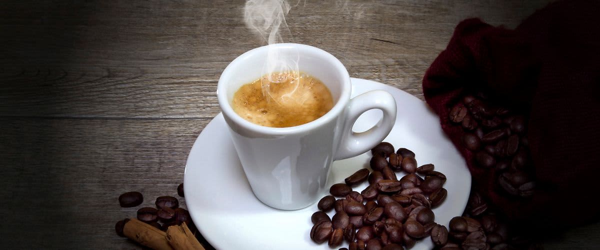 Šálek kávy na talířku obklopený kávovými zrny a skořicí na dřevěném stole