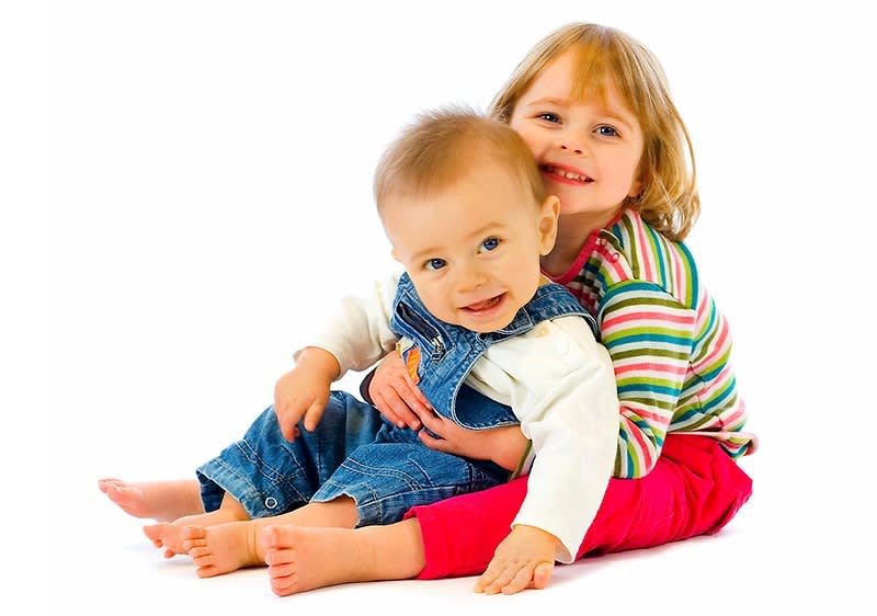 Dvě malé děti sedící na bílém pozadí, jedno v pruhovaném tričku, druhé v džínových lacláčích