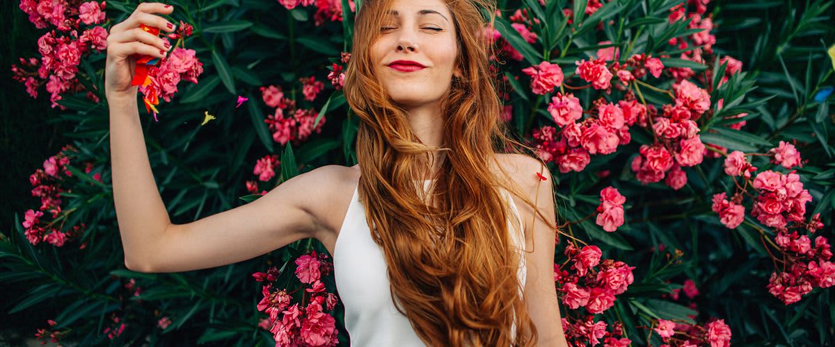 Žena s dlouhými zrzavými vlasy stojí před keřem s růžovými květy