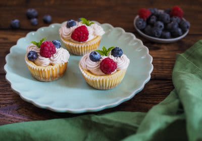 Tři cupcakes s krémem, malinami a borůvkami na modrém talíři, vedle mísa s ovocem a zelený ubrus