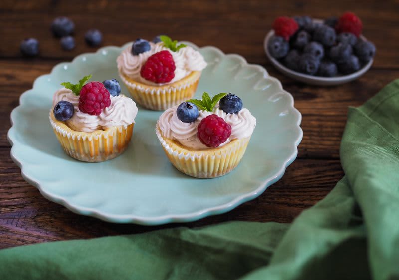 Tři cupcakes s krémem, malinami a borůvkami na modrém talíři, vedle mísa s ovocem a zelený ubrus