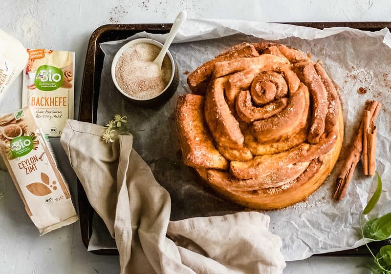 Pečená skořicová roláda na plechu s balíčky droždí a skořice, vedle misky s cukrem.