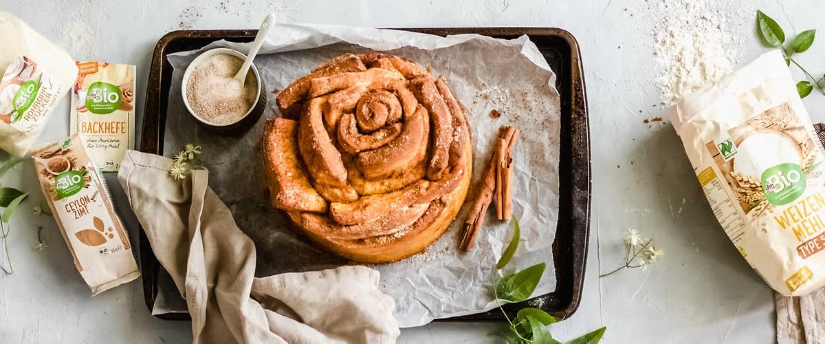 Pečená skořicová roláda na plechu s balíčky droždí a skořice, vedle misky s cukrem.
