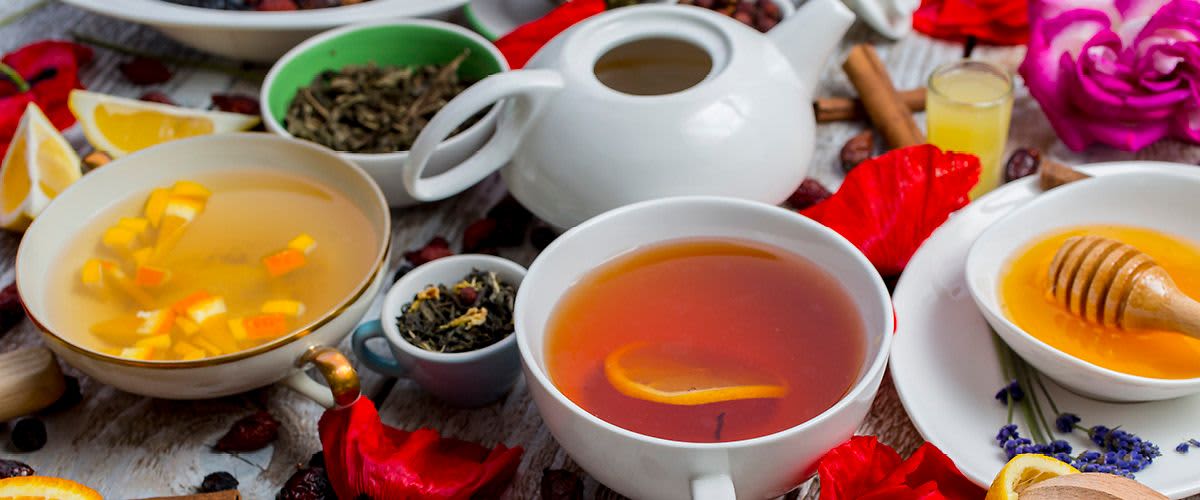 Různé druhy čaje v šálcích a konvici, sypaný čaj v miskách, plátky ovoce a květiny kolem
