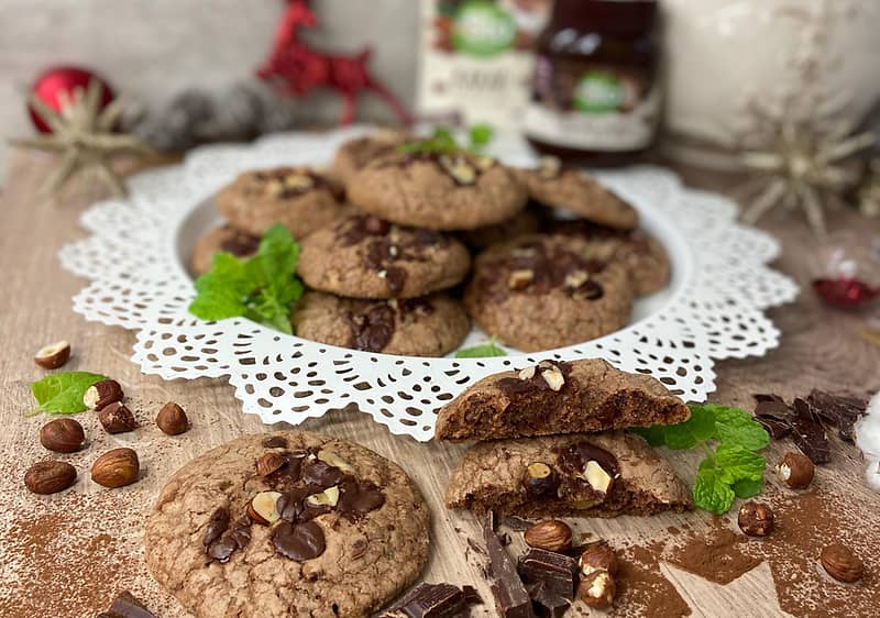 Čokoládové sušenky s oříšky na talíři, obklopené čokoládou a lístky máty na dřevěném stole.