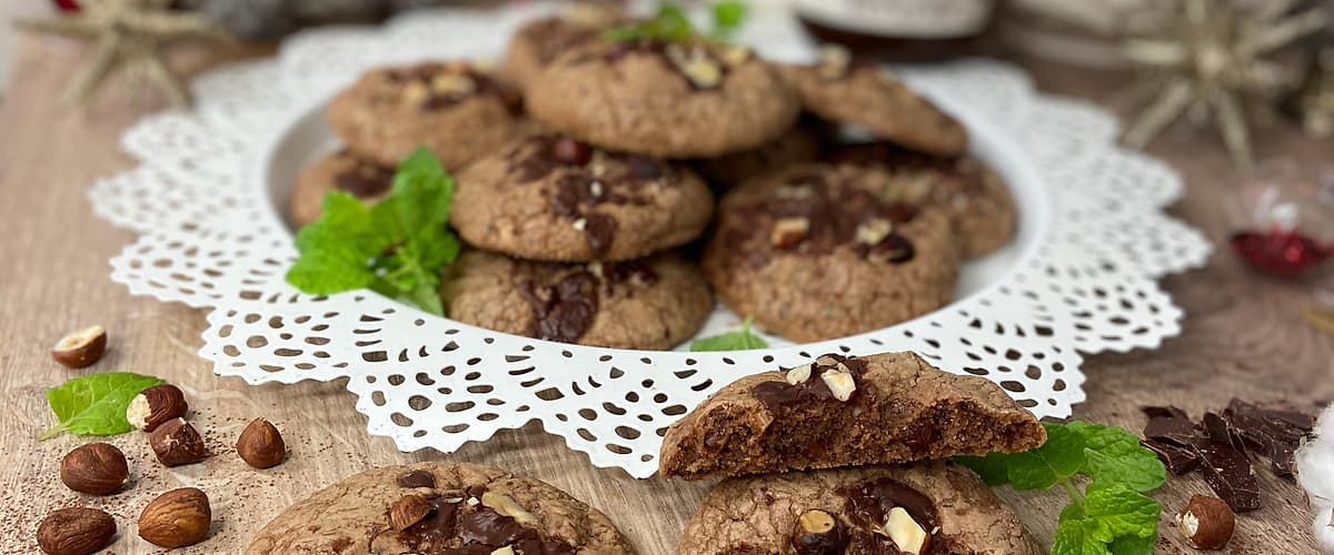 Čokoládové sušenky s oříšky na talíři, obklopené čokoládou a lístky máty na dřevěném stole.