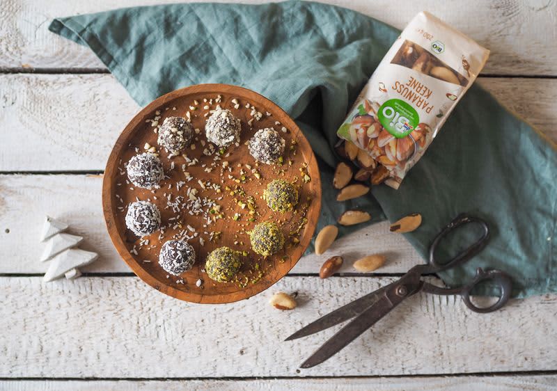 Dřevěný talíř s energetickými kuličkami, balení para ořechů, nůžky a zelený ubrousek na dřevěném stole