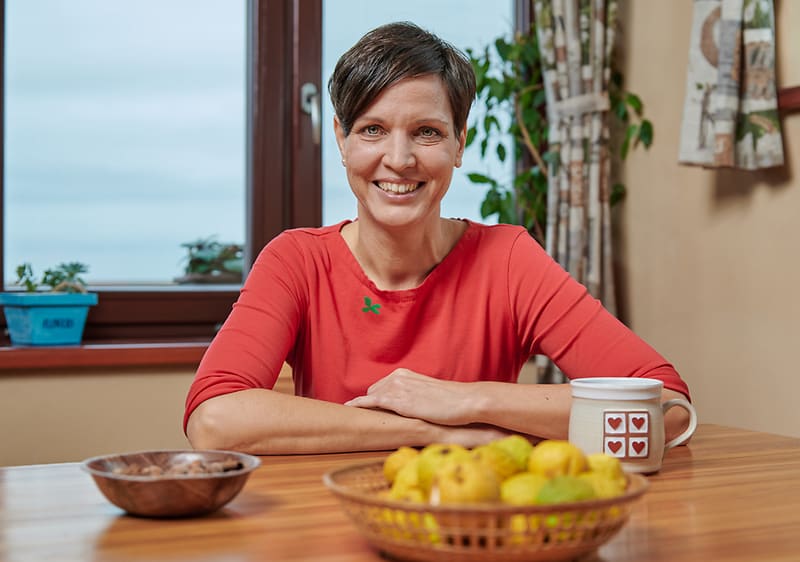 Žena u stolu s hrnkem, miskou a košíkem ovoce, rostliny na parapetu, závěsy a okno v pozadí