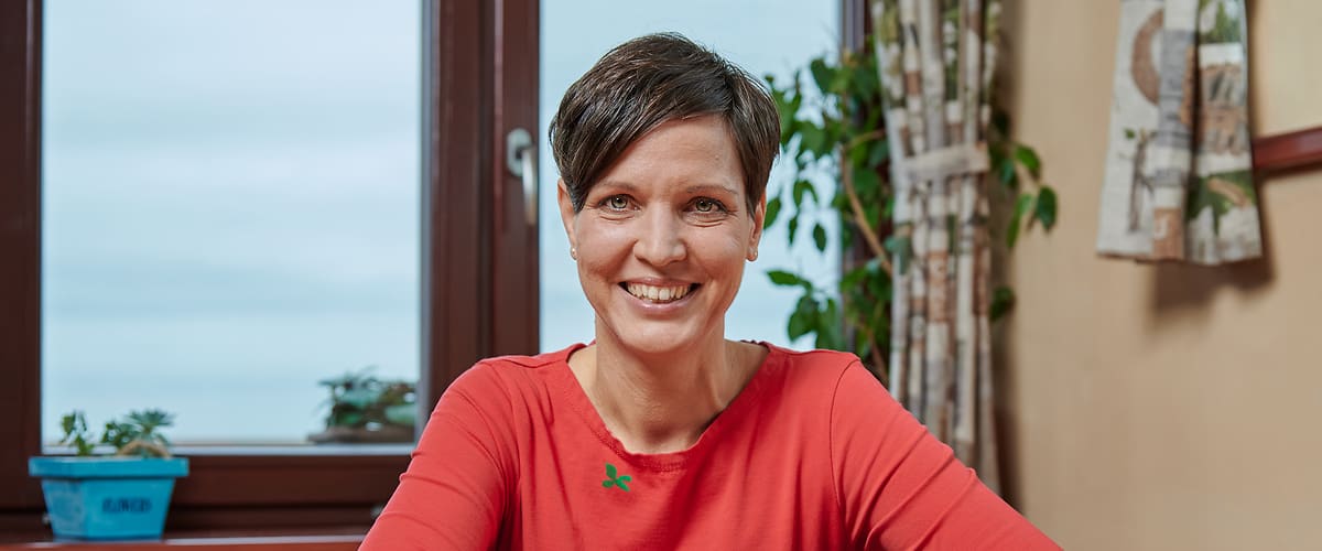 Žena u stolu s hrnkem, miskou a košíkem ovoce, rostliny na parapetu, závěsy a okno v pozadí