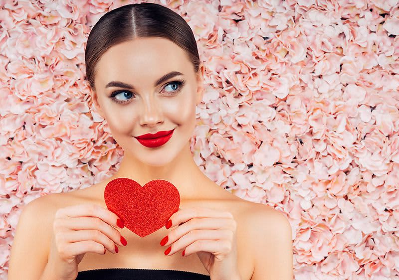 Žena držící červené srdce před květinovou stěnou z růžových květů