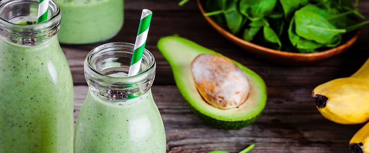 Avokádové smoothie v lahvi se zeleným brčkem, půlka avokáda a listy špenátu na dřevěném stole