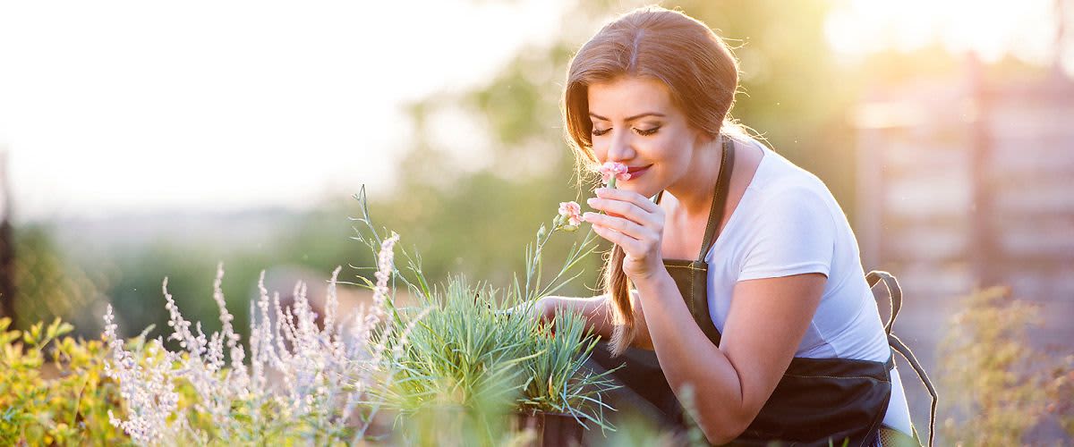 Žena v zahradě drží květinu, obklopená zelení a slunečním světlem.