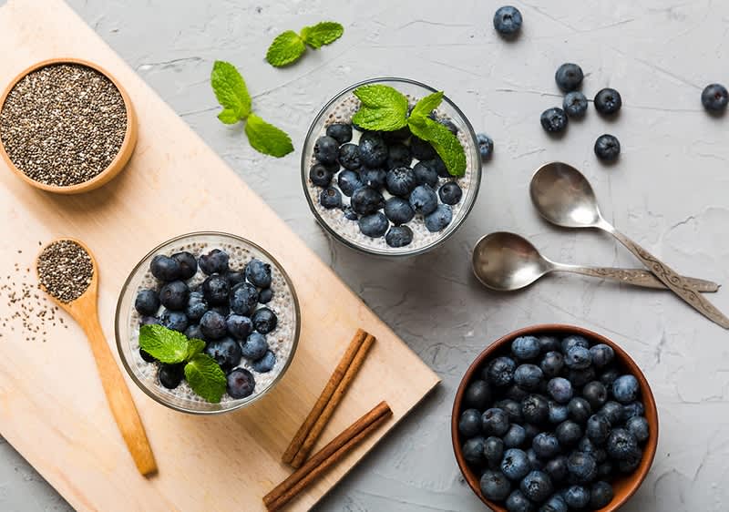 Chia pudink s borůvkami, mátou a skořicí na dřevěném prkénku, vedle misky s borůvkami a dvěma lžícemi.