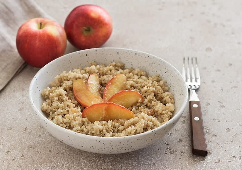 Miska s quinoou ozdobená plátky jablek, vedle dvě červená jablka a vidlička na stole.