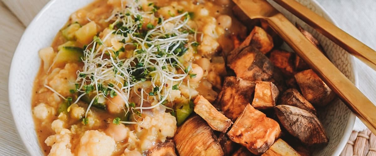 Miska s kari z cizrny a květáku, posypaná klíčky, vedle pečené batáty, zlatý příbor na straně.