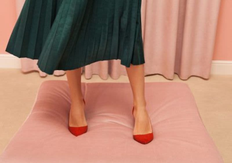 Žena v zelené sukni stojí na růžovém polštáři, má červené boty.