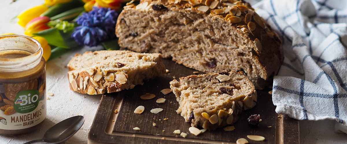 Chléb s mandlemi na prkénku, květiny, sklenice mandlového másla, lžíce a utěrka na stole