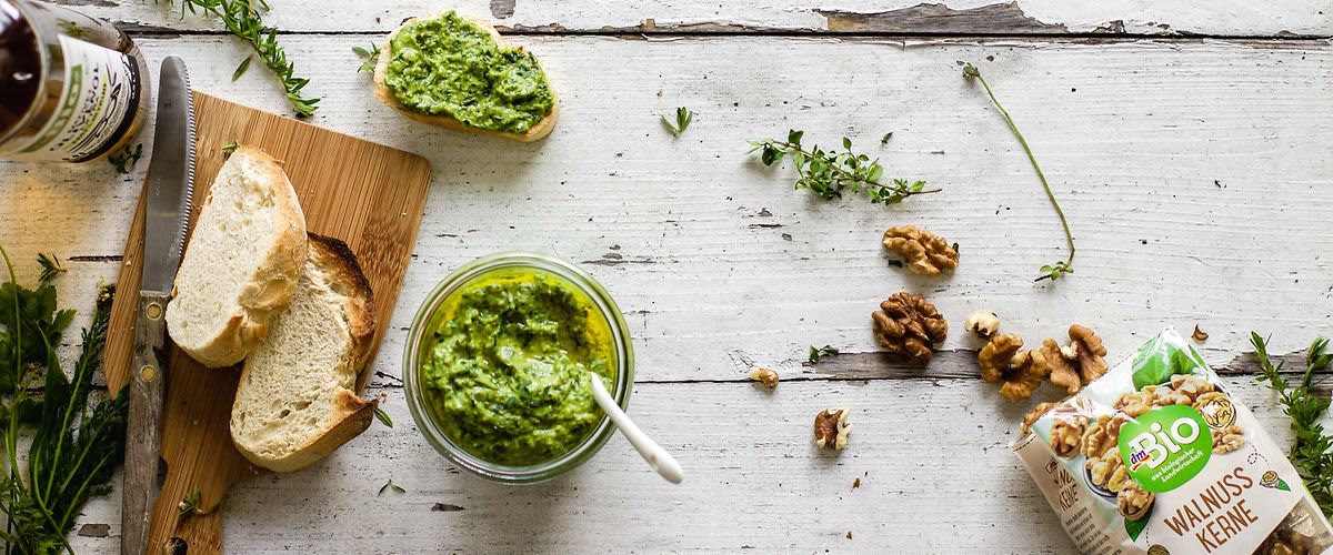 Chléb s pestem na prkénku, nůž, sklenice s pestem, láhev oleje, zelené bylinky