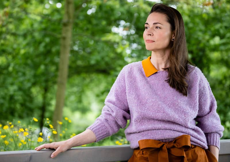 Žena v fialovém svetru a hnědé sukni stojí venku před zeleným pozadím