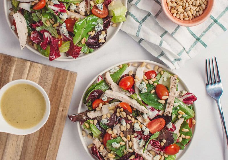 Salát s kuřecím masem, rajčaty, sýrem a piniovými oříšky na talíři, vedle dresink a vidlička