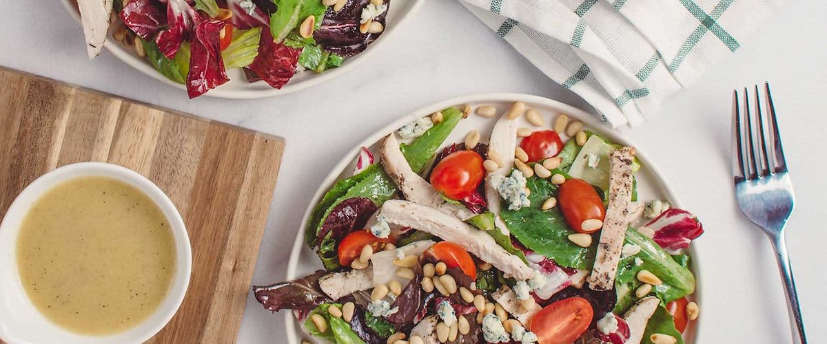 Salát s kuřecím masem, rajčaty, sýrem a piniovými oříšky na talíři, vedle dresink a vidlička