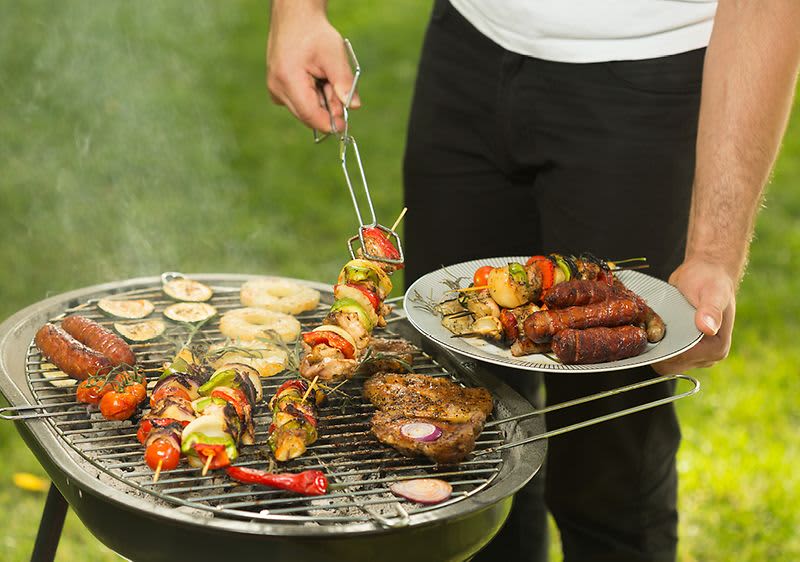 Grilování na zahradě s masem a zeleninovými špízy na grilu, muž drží talíř s hotovými pokrmy