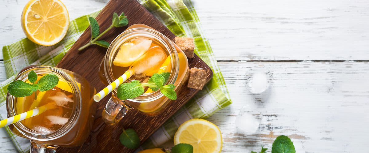 Ledový čaj s citronem a mátou ve sklenicích na dřevěném prkénku, obklopený citrony a kostkami cukru.