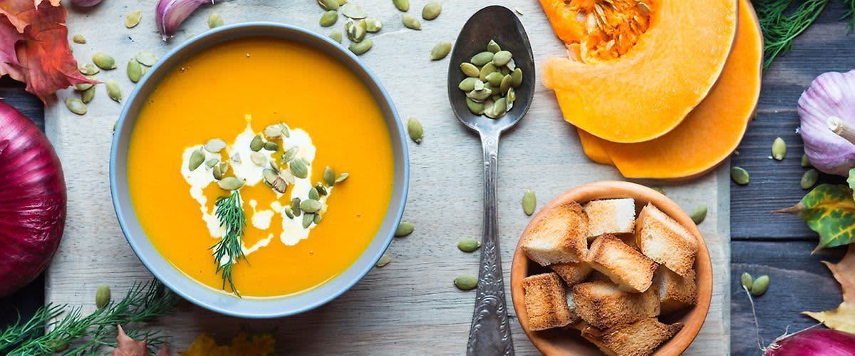 Dýňová polévka s dýňovými semínky, smetanou a koprem, vedle lžíce a misky s krutony