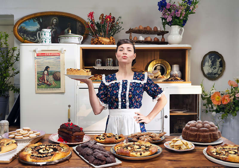 Žena v kuchyni s dortem, stůl plný různých dezertů, květiny a starožitný nábytek v pozadí