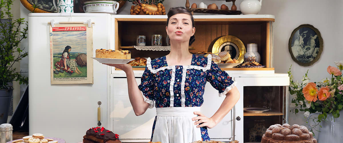 Žena v kuchyni s dortem, stůl plný různých dezertů, květiny a starožitný nábytek v pozadí