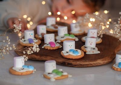 Dítě pozoruje sušenky s marshmallows a barevnými bonbony na dřevěném prkénku, ozdobené světýlky.