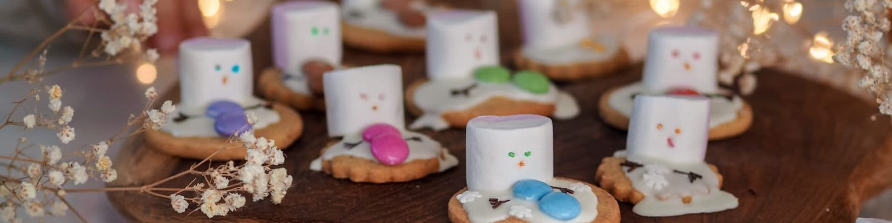 Dítě pozoruje sušenky s marshmallows a barevnými bonbony na dřevěném prkénku, ozdobené světýlky.